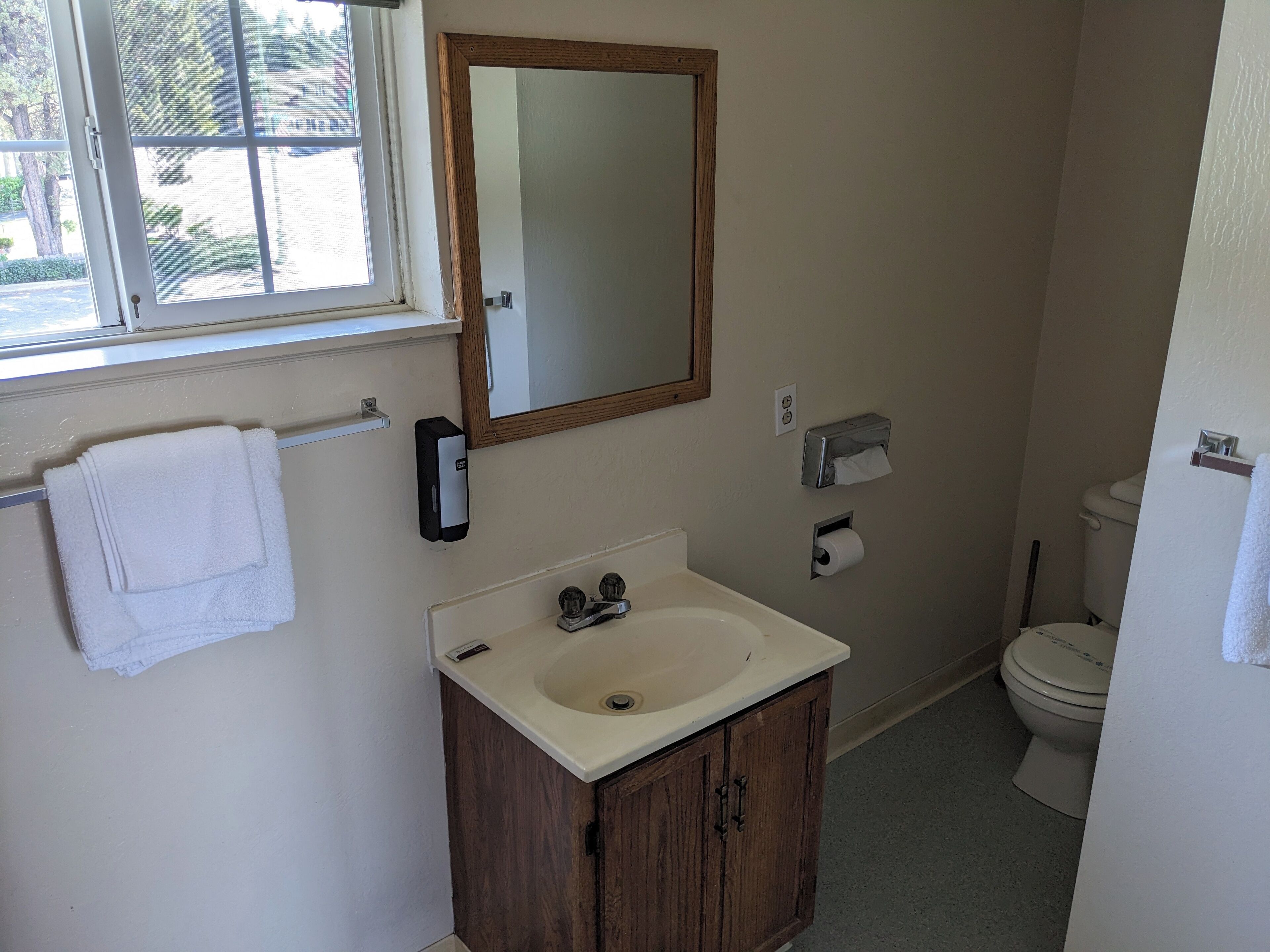 Une salle de bain avec un meuble en bois, un lavabo, un miroir, un porte-serviettes, des toilettes et une fenêtre.