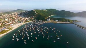 Vista aérea de un pueblo costero con un puerto deportivo lleno de barcos, una playa y montañas al fondo.