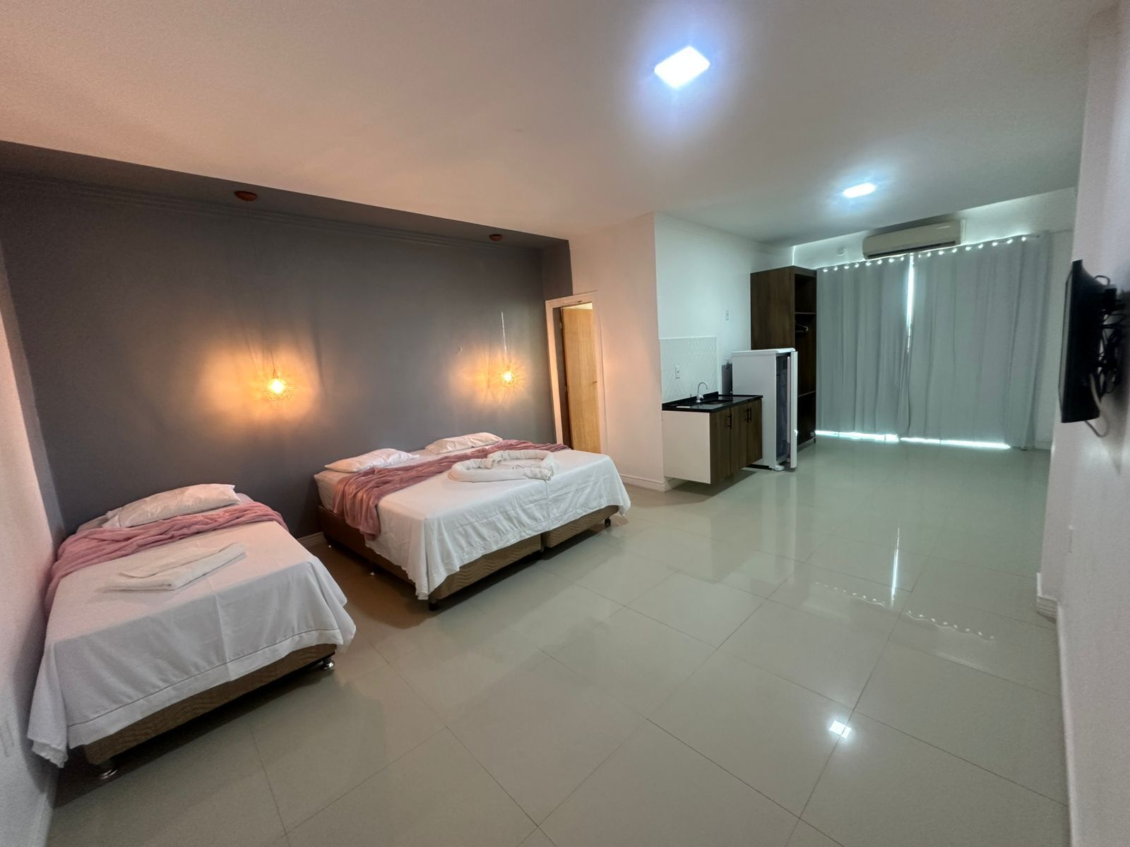 A hotel room with two beds, a kitchenette, a mirror, and a sliding door leading to a balcony.