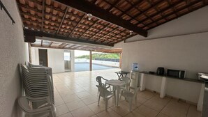Una habitación amplia con suelo de baldosas, techo de madera, zona de comedor con sillas de plástico y una mesa, cocina con microondas y dispensador de agua, y vista a una piscina a través de un vano abierto.