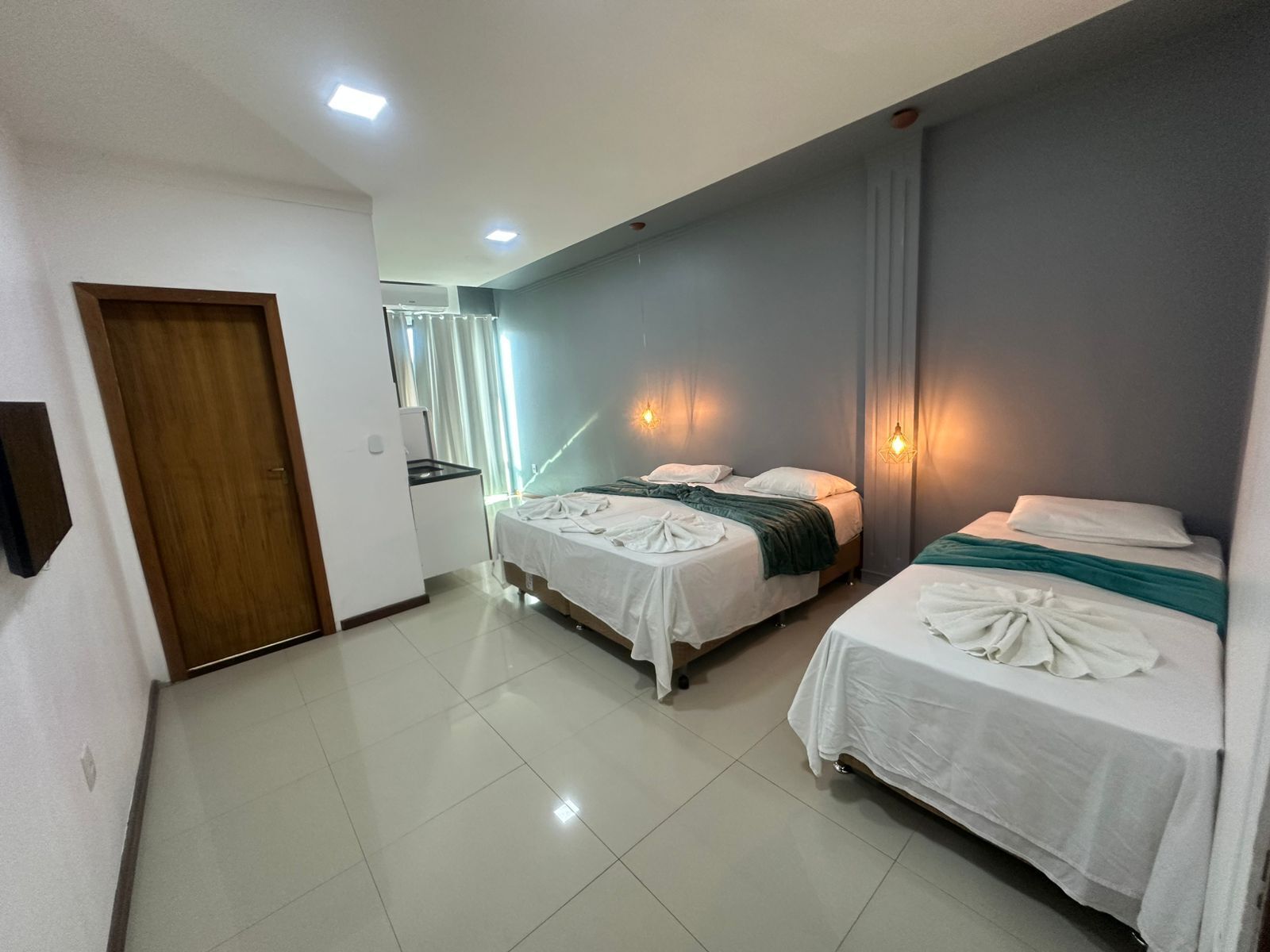Habitación de hotel con dos camas, un pequeño kitchenette y un baño visible a través de una puerta de vidrio.