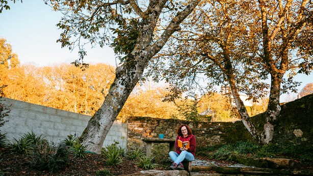 A person sitting on a wooden bench in a garden with trees and a stone wall.