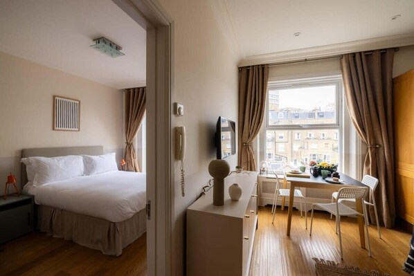 A hotel room with a bed, a desk, a chair, and a window with curtains. - The Courtfield - 1 Bed Apartment in Kensington (London)