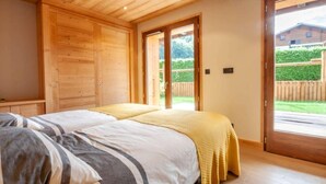 A bedroom with two beds, wooden walls, a large sliding door to a balcony, and a view of a house and greenery.