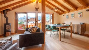 Ein großzügiger Wohnbereich mit Holzbalkendecke, einer Couch, einem Esstisch mit Stühlen und Blick auf eine Berglandschaft durch große Fenster.