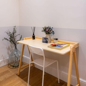 Un petit bureau en bois, une chaise blanche, un vase avec des fleurs, un livre et un petit panneau.