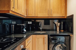 A kitchen with wooden cabinets, a black countertop, a coffee maker, a kettle, a sink, and a washing machine. - The Courtfield - Studio Apartment in Kensington (London)