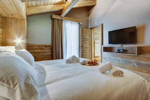 Hotellihuone, jossa on puinen katto, valkoisilla lakanoiden peitetty sänky, taulutelevisio ja aamiaistarjoilu sängyn päällä.