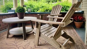 Una silla Adirondack de madera y una mesa redonda en un porche con pared de ladrillo, plantas en macetas y vista a una casa y follaje.