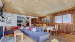 A cozy living room with a grey sofa, wooden dining table, and a staircase leading to an upper floor. - Chalet, 70m from ski lifts, view mountain, swimming pool, sauna, fireplace or stove, parking, tv (Morzine)