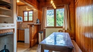 A wooden cabin interior with a kitchen area, a dining table, and a window with a view of the forest.