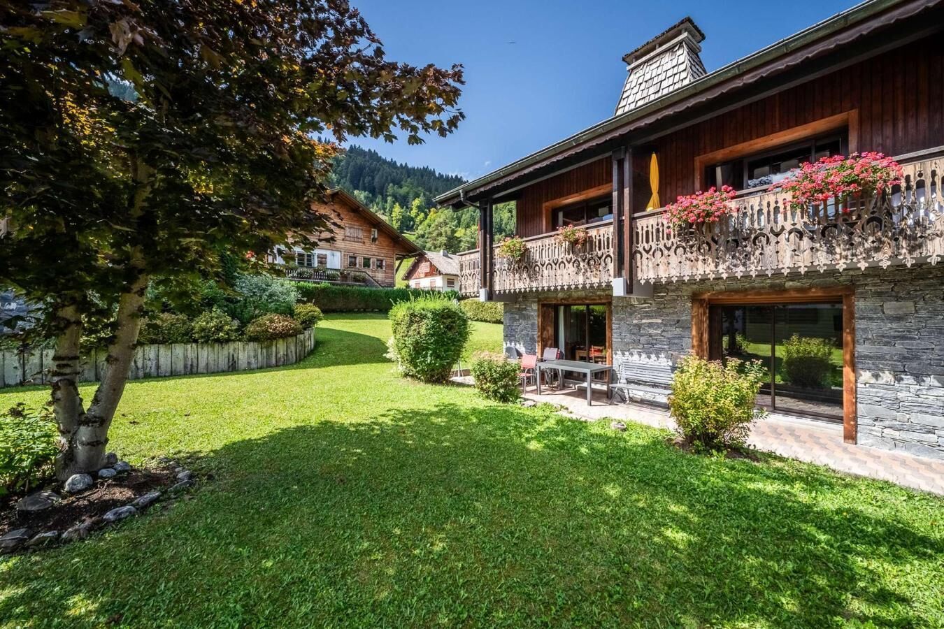 A two-story chalet with a stone facade, a balcony with flowers, and a well-maintained garden.