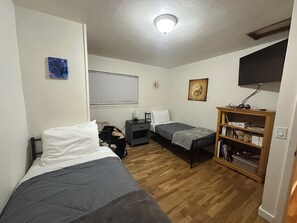 Ein kleiner, gut beleuchteter Raum mit zwei Einzelbetten, einem Nachttisch mit Lampe, einem Holzregal und einem Fenster mit Jalousien.