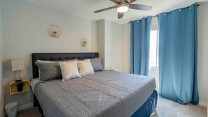 A bedroom with a bed, a ceiling fan, a window with curtains, and a nightstand with a lamp.