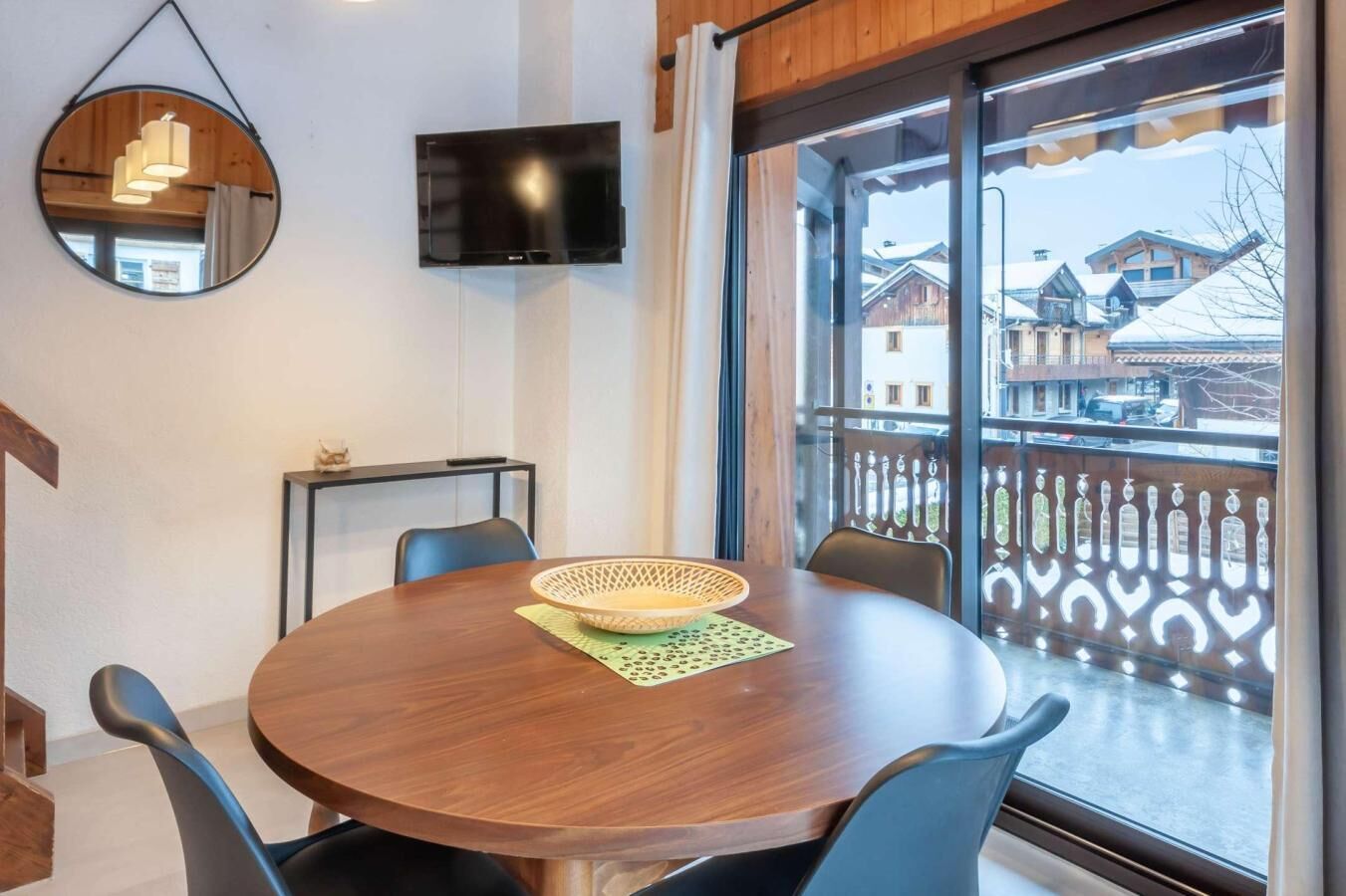 A round wooden table with chairs, a television mounted on the wall, and a view of snow-covered houses through large windows.