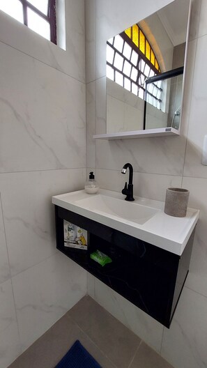 A bathroom with a white sink, black faucet, and a mirror above it. There is a towel dispenser and a toilet paper holder on the wall.