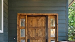 Puerta de madera con paneles de vidrio y un manillar de metal, ubicada en un porche con revestimiento exterior oscuro.