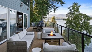 Una terraza con un sofá, sillas, una fogata y una mesa, con vistas a un lago, árboles y montañas al fondo.