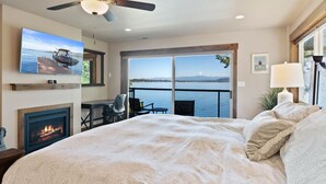 Un dormitorio con una cama grande, ventilador de techo, chimenea, televisor y balcón con vista al agua.