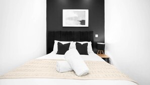 A neatly made bed with white and black bedding, a rolled white towel, and a small wooden side table with a chair.