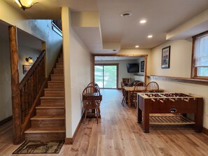 A wooden-floored room with a foosball table, a dining area with chairs, a staircase, and a television mounted on the wall.