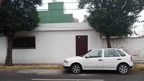 Un coche blanco estacionado en una calle con un edificio blanco que tiene una puerta marrón y una ventana con una celosía metálica.