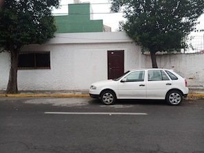 A white car parked on a street with a white building featuring a brown door and a window with a metal grille. - Apartamento Céntrico Vive El Pulsar De La Ciudad (Toluca)