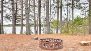 Un campamento rústico con un brasero metálico rodeado de pinos y un lago al fondo.