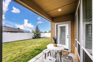 A patio with a table and chairs, a door, and a view of a backyard with a lawn and a white fence.