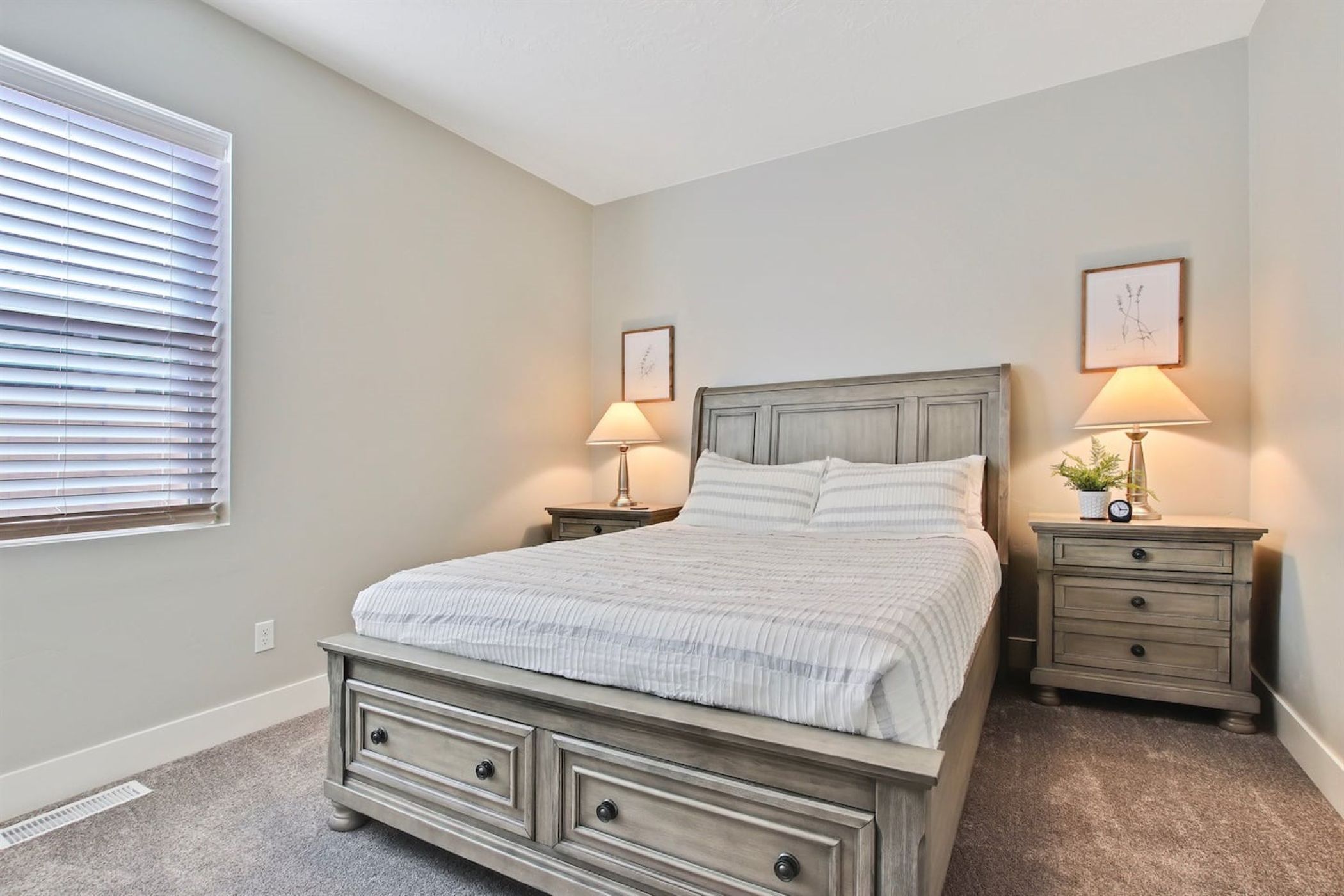 A bedroom with a bed, two nightstands, a dresser, a window with blinds, and wall art.