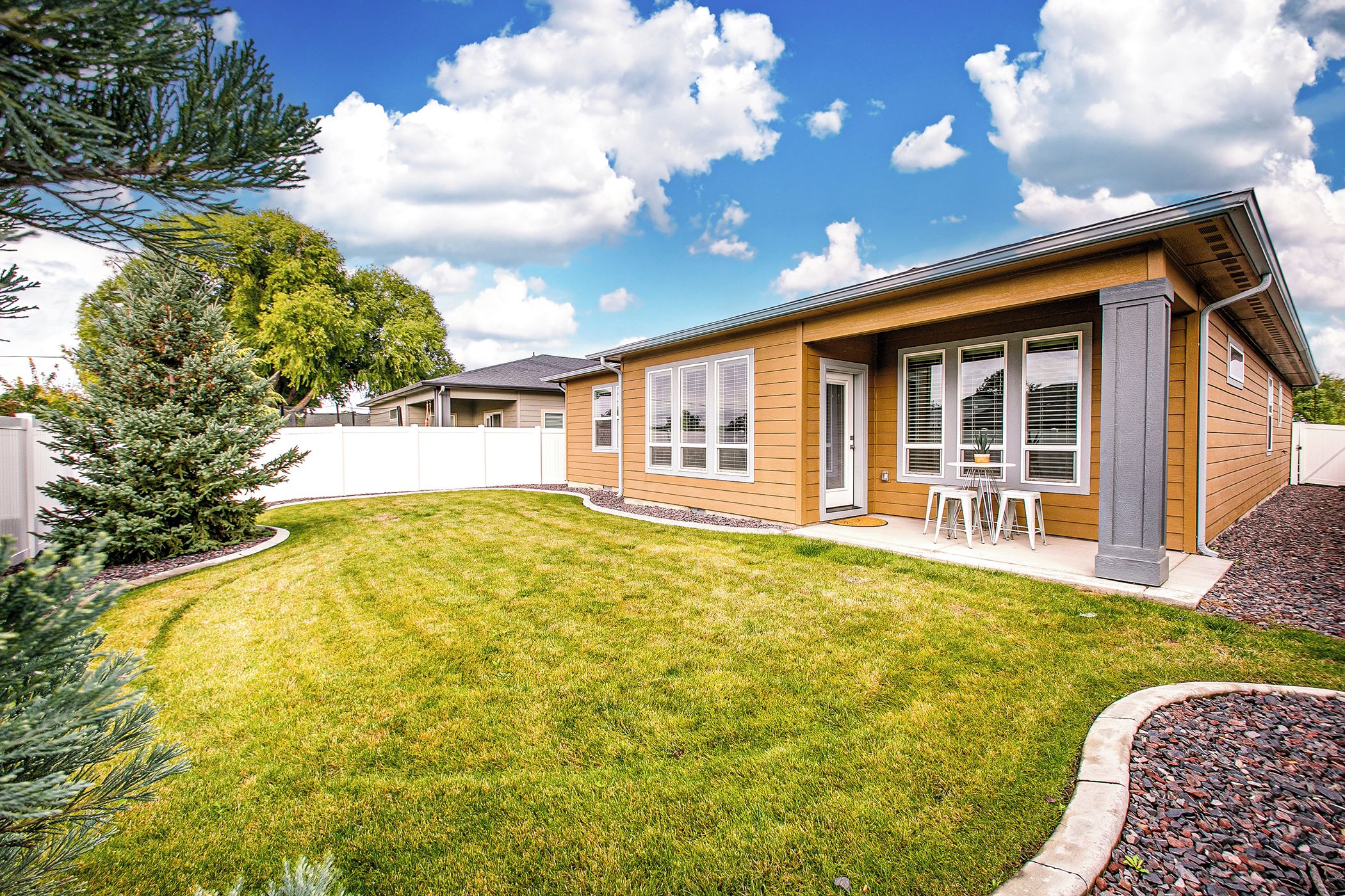 A modern single-story house with a well-maintained lawn, a patio with outdoor furniture, and a large window on the front.