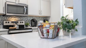 A modern kitchen with white cabinets, a stainless steel microwave, and a kettle on the stove. There is a wire basket filled with groceries on the counter. - Newly Renovated Townhouse! (Davenport)