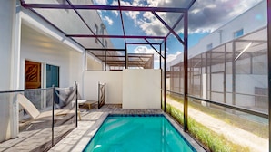 Uma piscina com área coberta, um caminho de tijolos e vista para um edifício ao fundo.