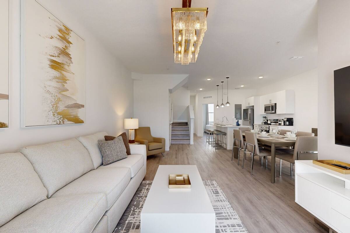 A modern living room with a sofa, coffee table, and a chandelier. There is a dining area with chairs and a table, and a kitchen with appliances.
