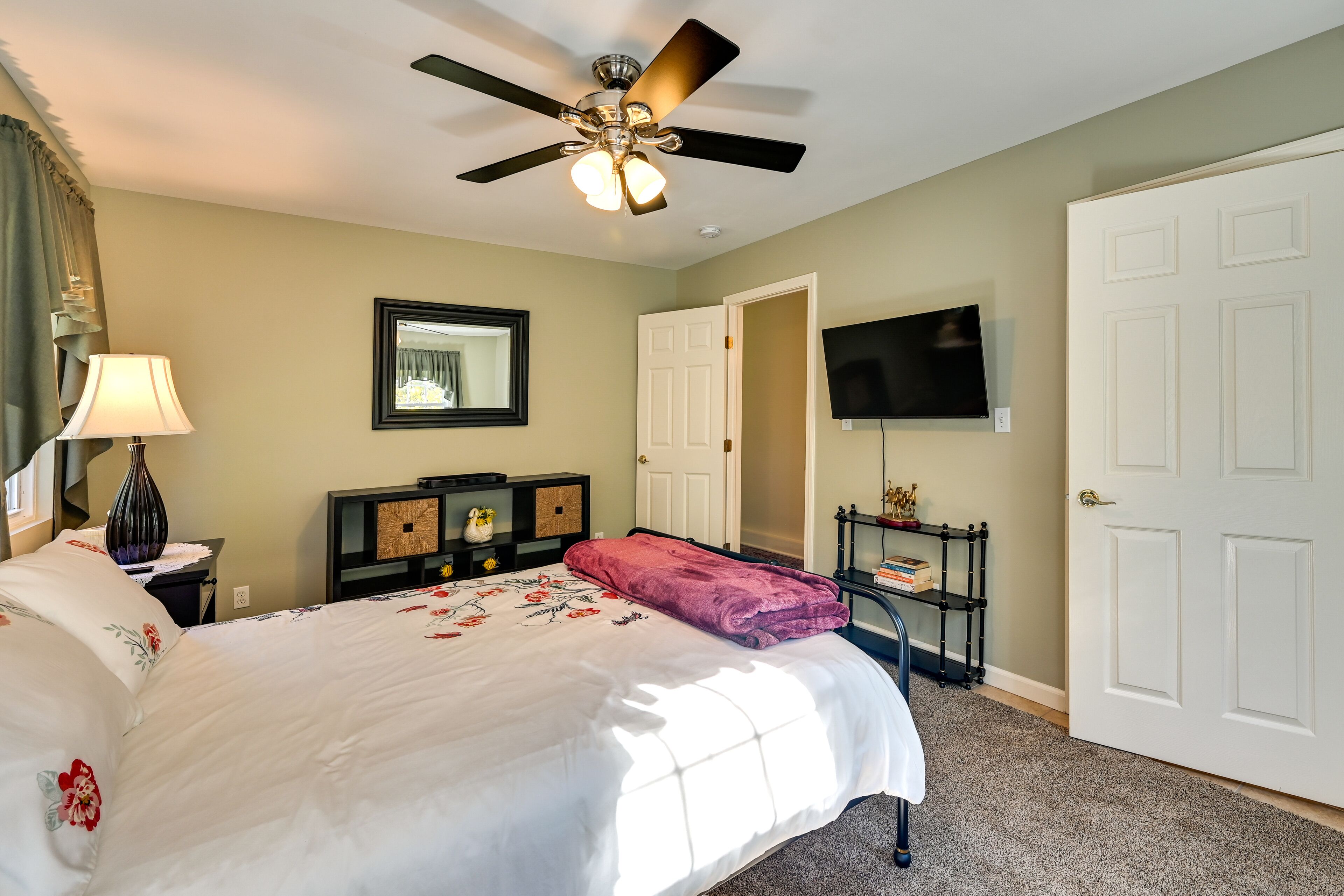 A hotel room with a bed, ceiling fan, TV, and a door.