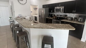 A modern kitchen with dark cabinets, a granite countertop, a stainless steel refrigerator, and a microwave. There are three stools and a clock on the wall. - Come relax, restore and play at this calming fun retreat! Location! Pool, BBQ. (Cape Coral)