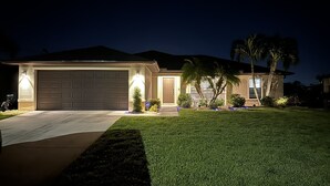 A residential house with a garage, well-lit entrance, and a lawn with palm trees. - Come relax, restore and play at this calming fun retreat! Location! Pool, BBQ. (Cape Coral)