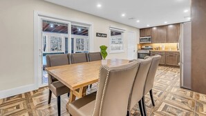 A dining area with a wooden table and beige upholstered chairs, a kitchen with dark cabinets, and a view through large windows.
