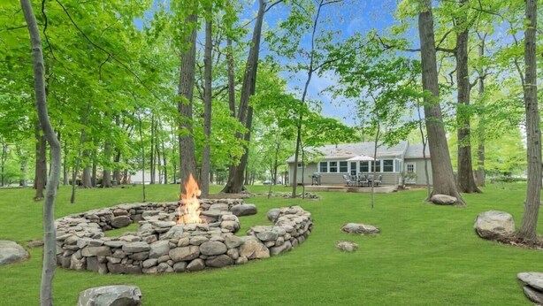 A stone fire pit surrounded by rocks, a grassy yard, and a house with a covered patio.
