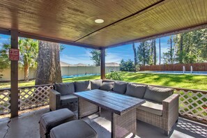 An outdoor patio with a wooden table and wicker sofa, overlooking a swimming pool and a grassy yard.