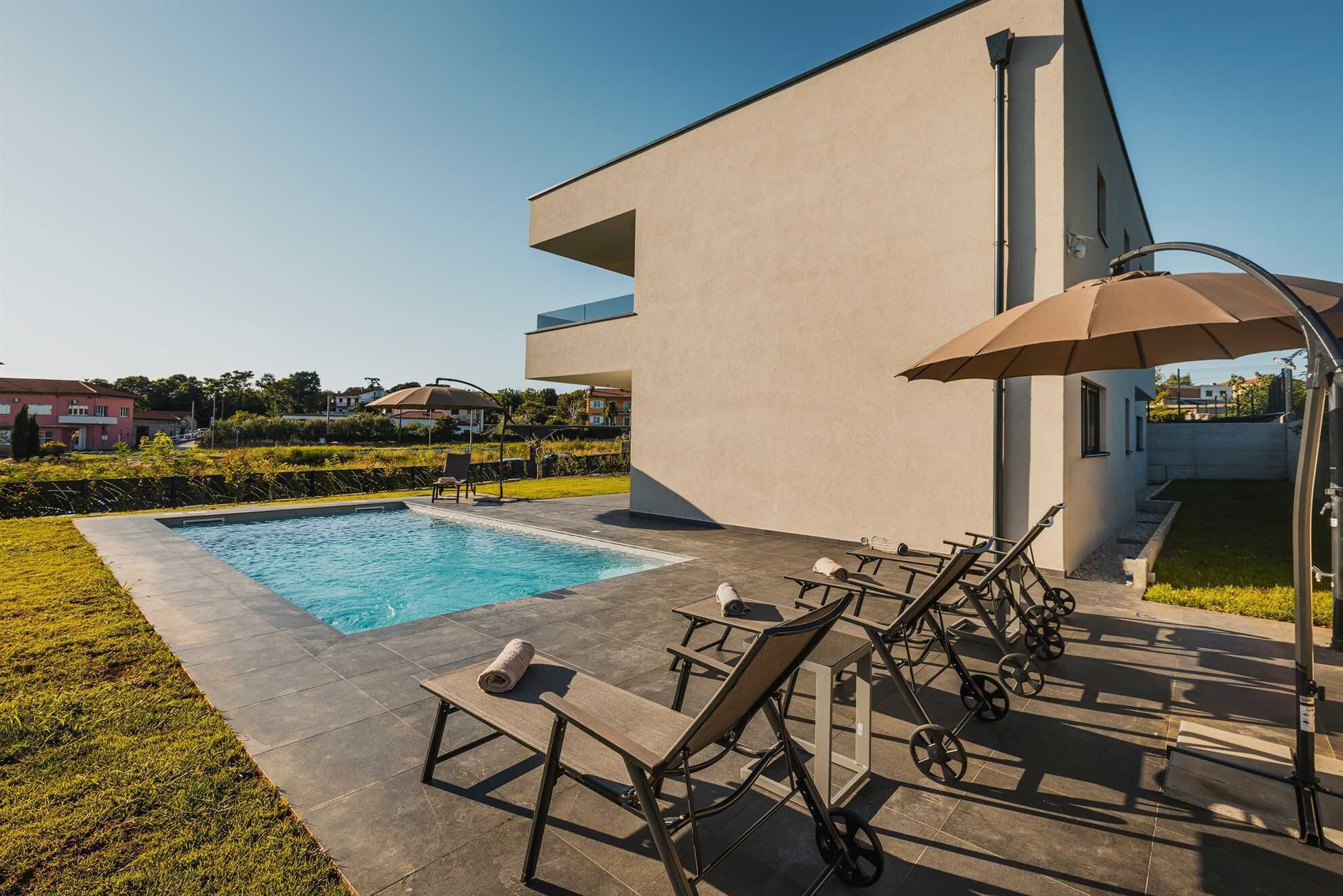 Un espace piscine extérieur moderne comprenant une piscine rectangulaire, des chaises longues et une table avec un parasol.