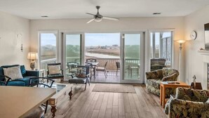 Een ruime woonkamer met een grote schuifpui naar een balkon, een plafondventilator en een eethoek met een tafel en stoelen.