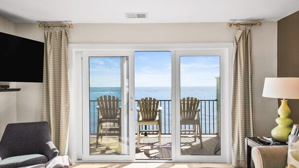 Een kamer met een grote schuifglazen deur die leidt naar een terras met uitzicht op de oceaan. Op het terras staan twee houten stoelen, een televisie is aan de muur bevestigd en op een bijzettafeltje staat een lamp.