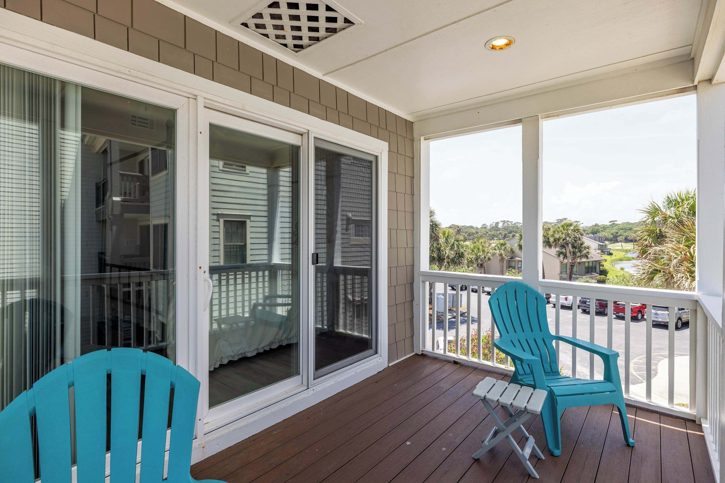 A covered porch with a view of parked cars and palm trees, two blue Adirondack chairs, and a sliding glass door.