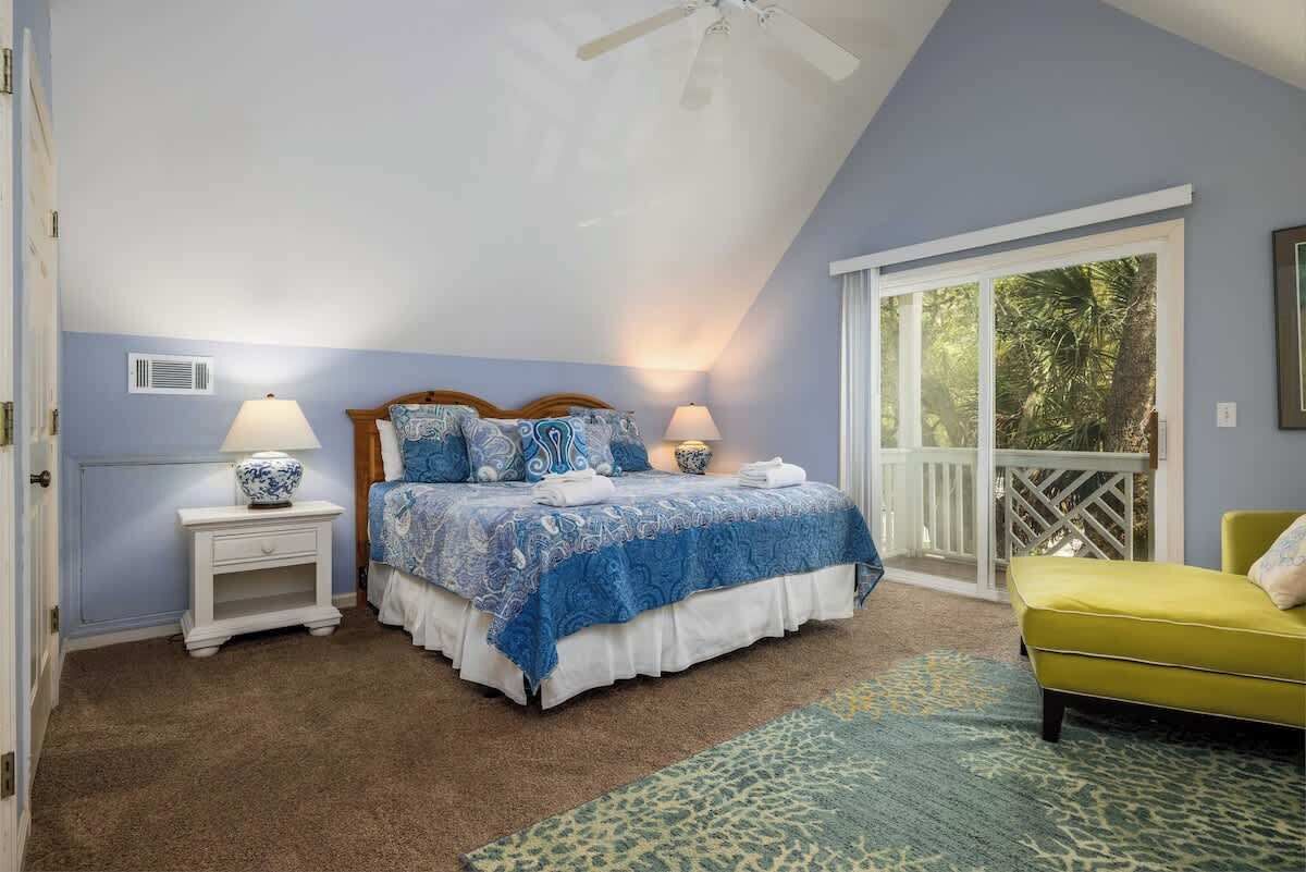 A bedroom with a bed, two bedside tables with lamps, a window with a view, a balcony, and a yellow chair.