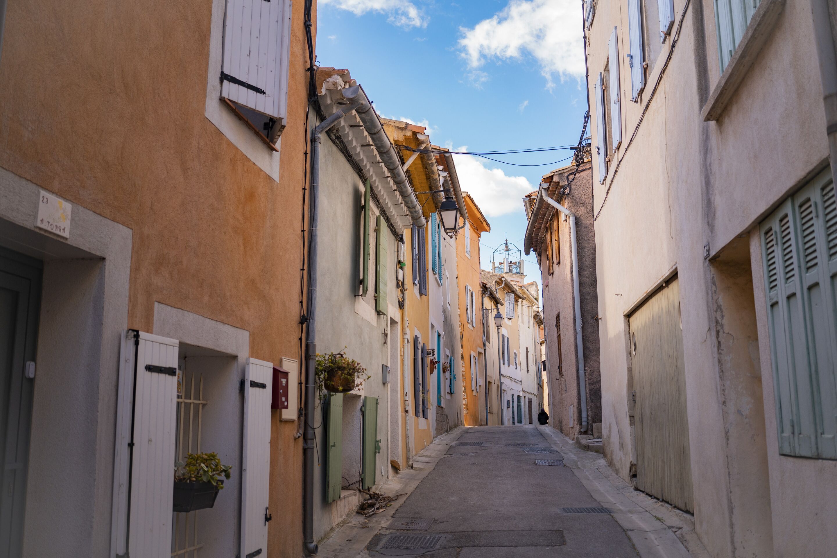 A narrow street with buildings on both sides, featuring traditional architecture with shutters and balconies.
