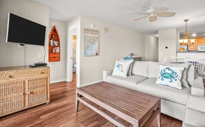 A living room with a television, a bookshelf, a white sofa with decorative pillows, a wooden coffee table, and a ceiling fan.