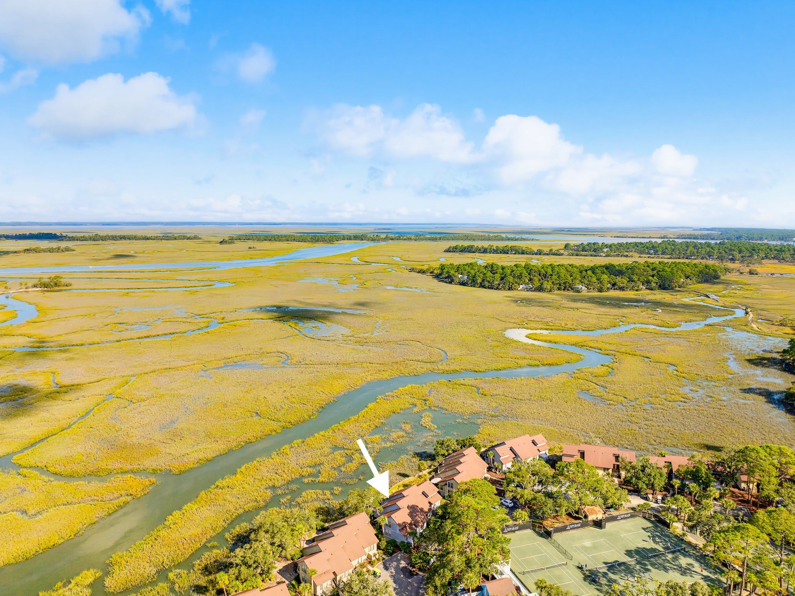 An aerial view of a coastal area with a river, marshland, and a residential complex with houses and a tennis court.