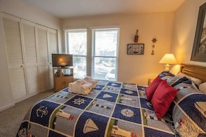 Quarto com uma cama grande, televisão em móvel, janela com persianas e armário com portas brancas.