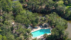 An aerial view of a resort with a swimming pool surrounded by dense trees and a small building with a roof. - 271 Deer Lake Drive (Fripp Island)